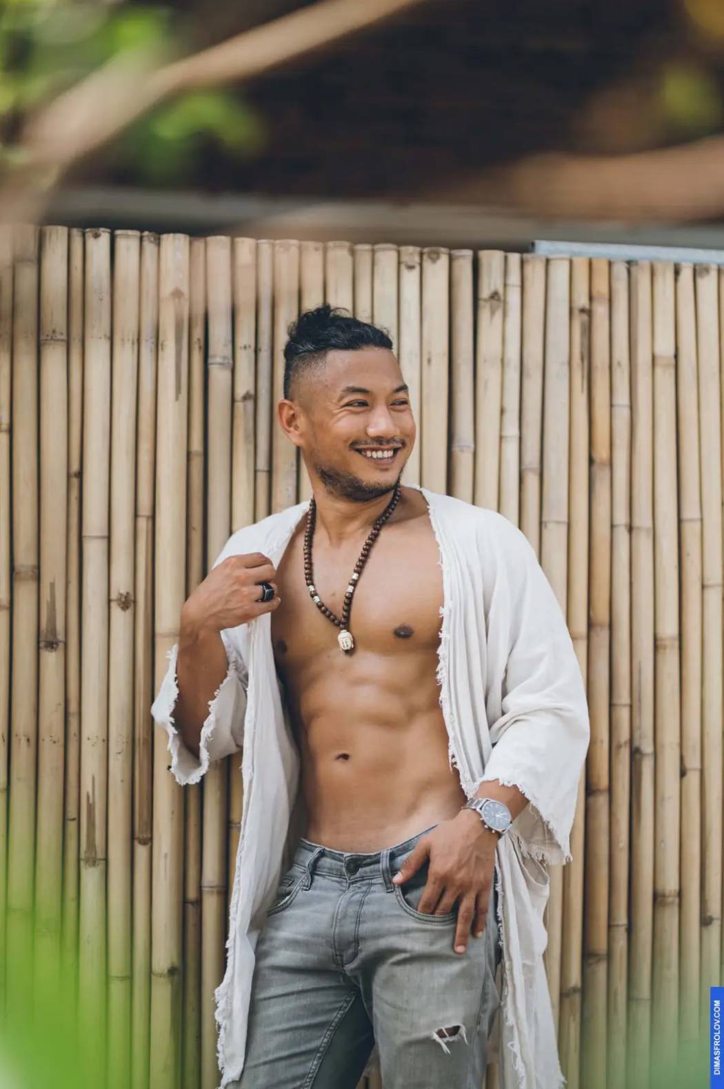 Smiling shirtless man in open white shirt showing abs in tropical setting