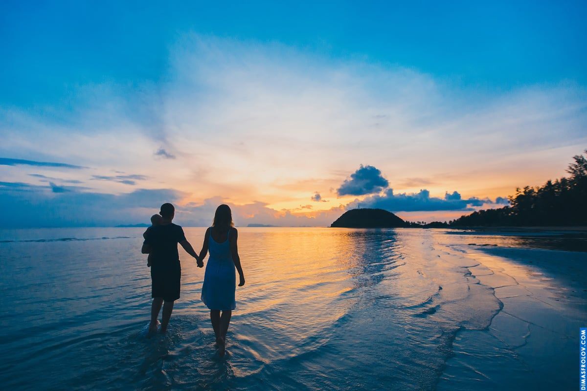 Sessão de fotos em família ao pôr do sol — pais caminhando em mar raso com o bebê nos braços do pai