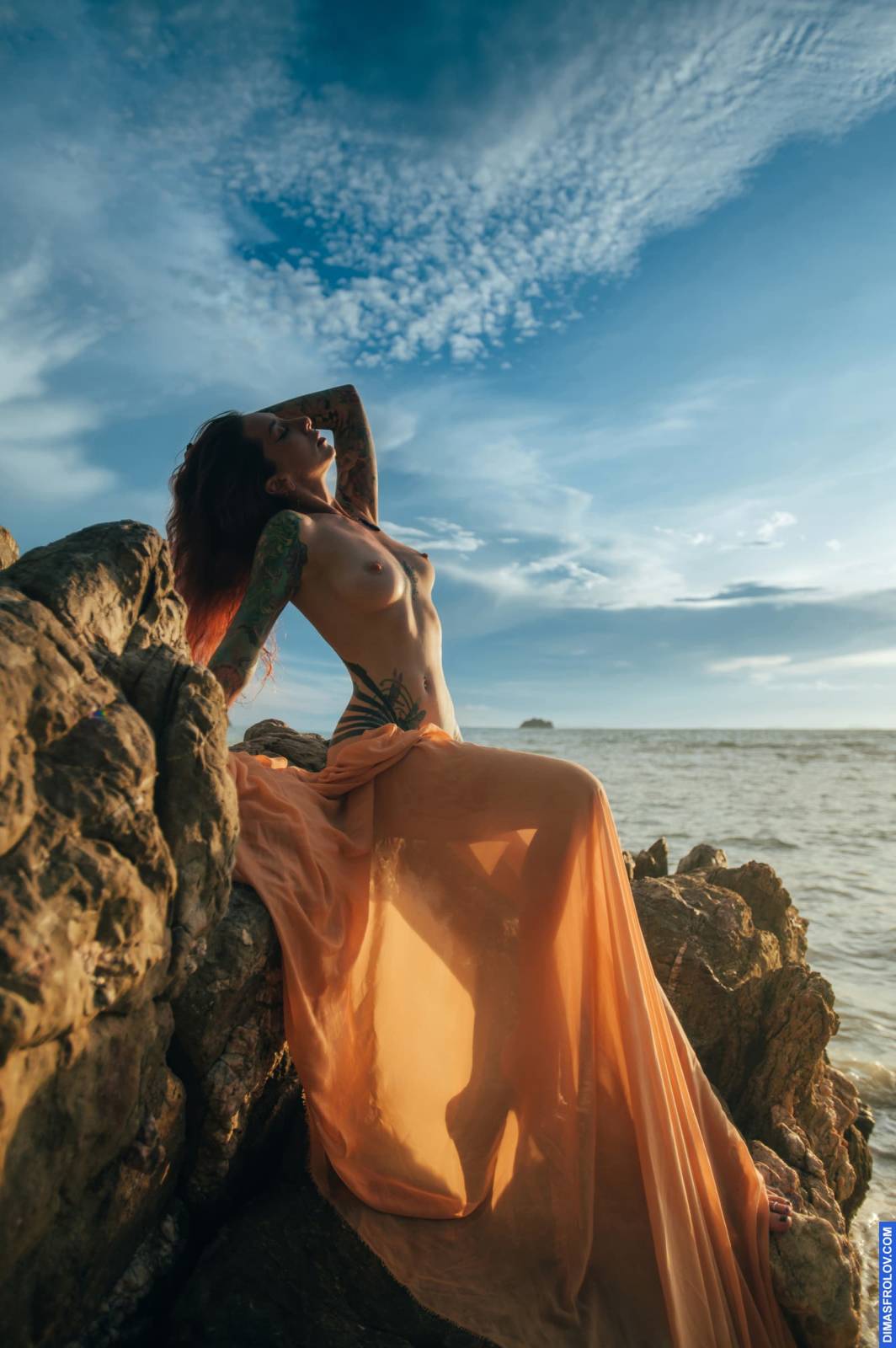 Retrato nu na hora dourada em rochas à beira-mar com saia transparente esvoaçante