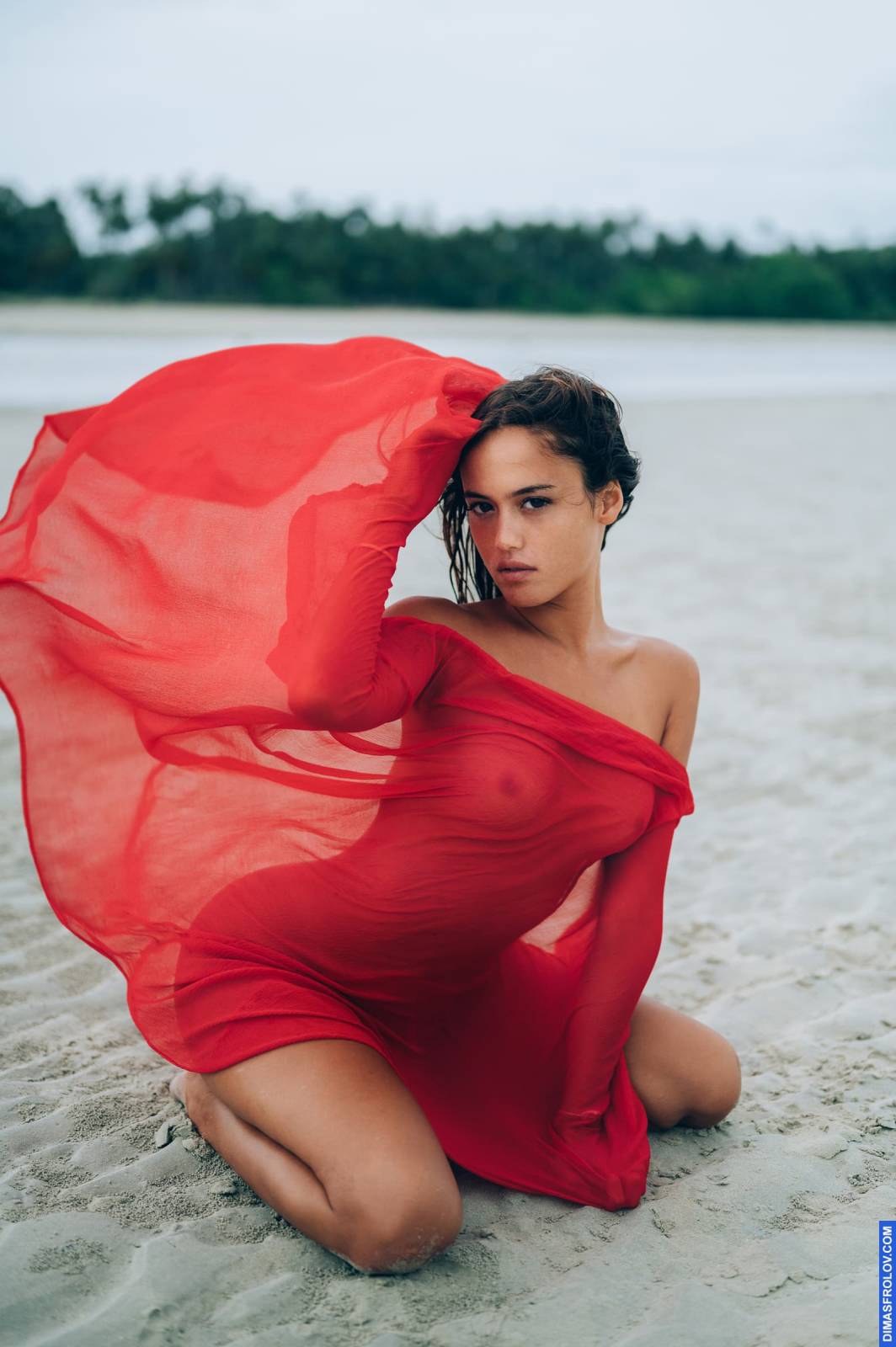 Retrato artístico nu de uma mulher envolta em um tecido vermelho transparente, ajoelhada na praia