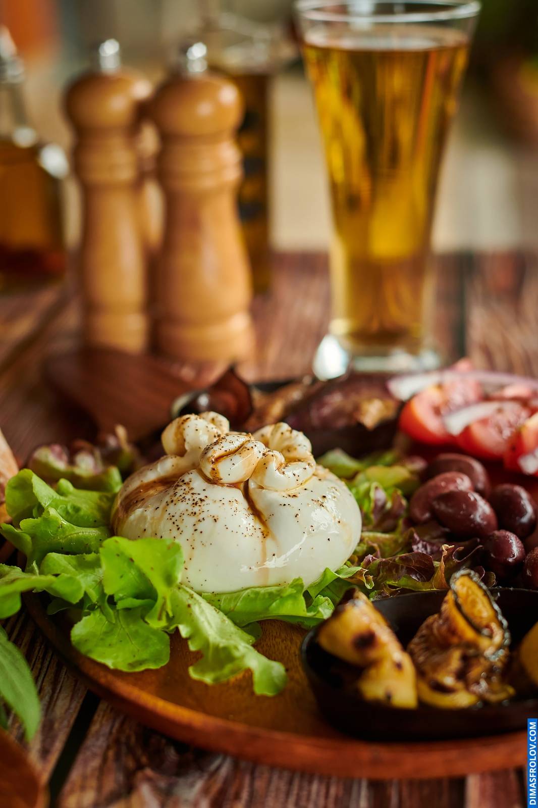 Life-style shot in natural light featuring a plate with burrata cheese, vegetables, and olives, served with beer, ideal for restaurants and food bloggers showcasing fresh and light Mediterranean cuisine