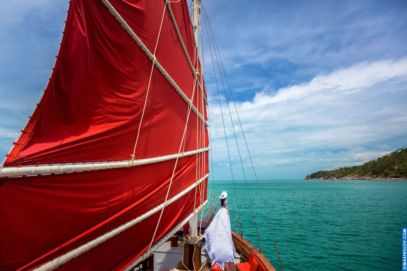 Woman in a flowing white dress posing on a red sailboat during a fashion photo set