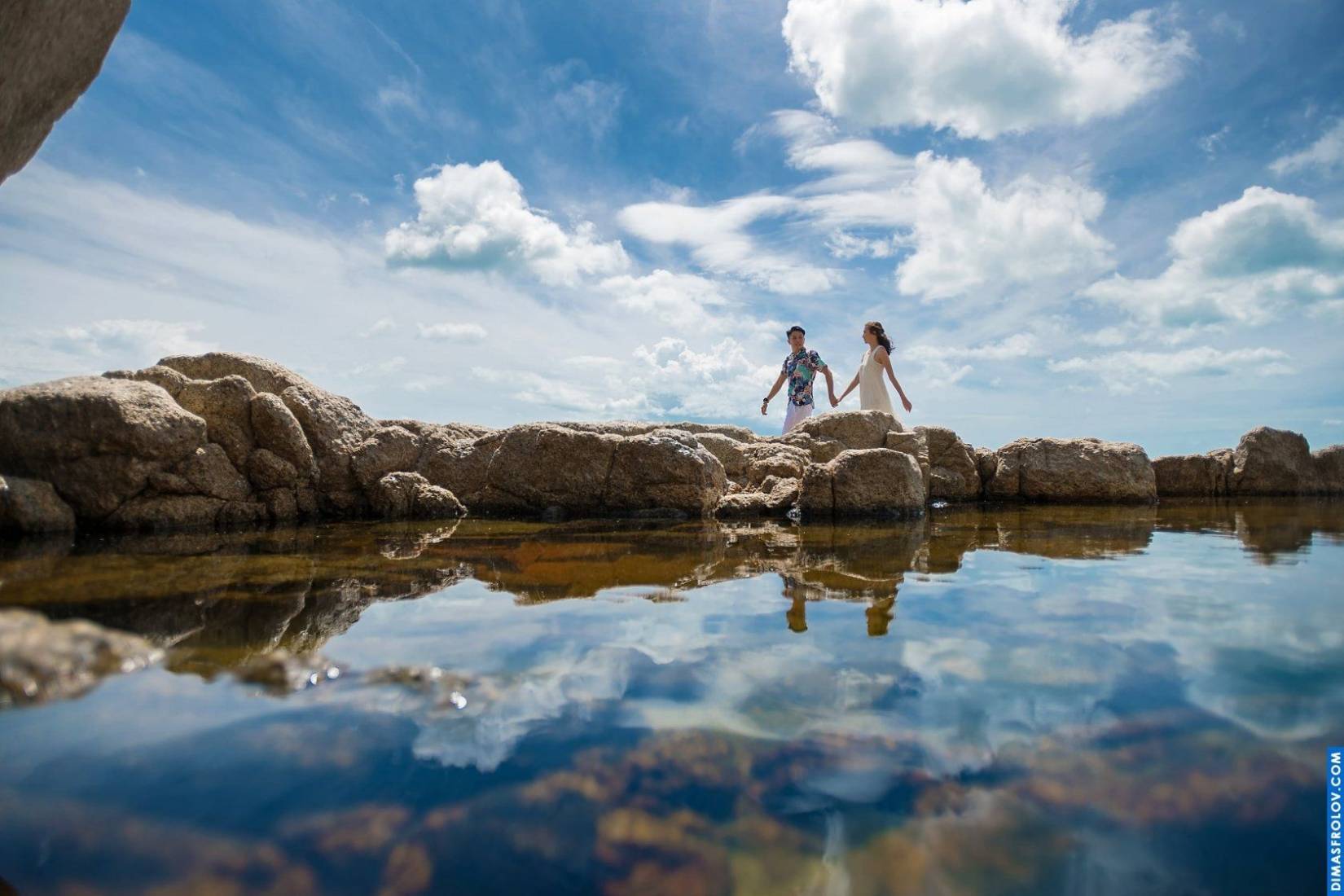 Engagement photo with a couple walking into the sunset through calm water, with mirrored clouds and dramatic wide-angle reflections