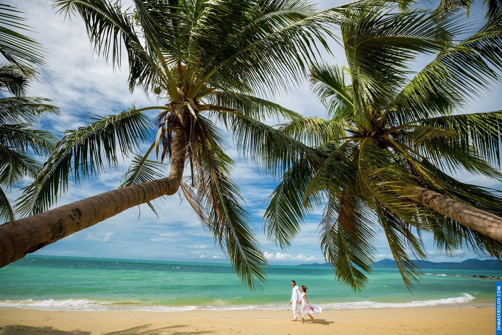 Couple in white walking along an empty tropical beach under palm trees
