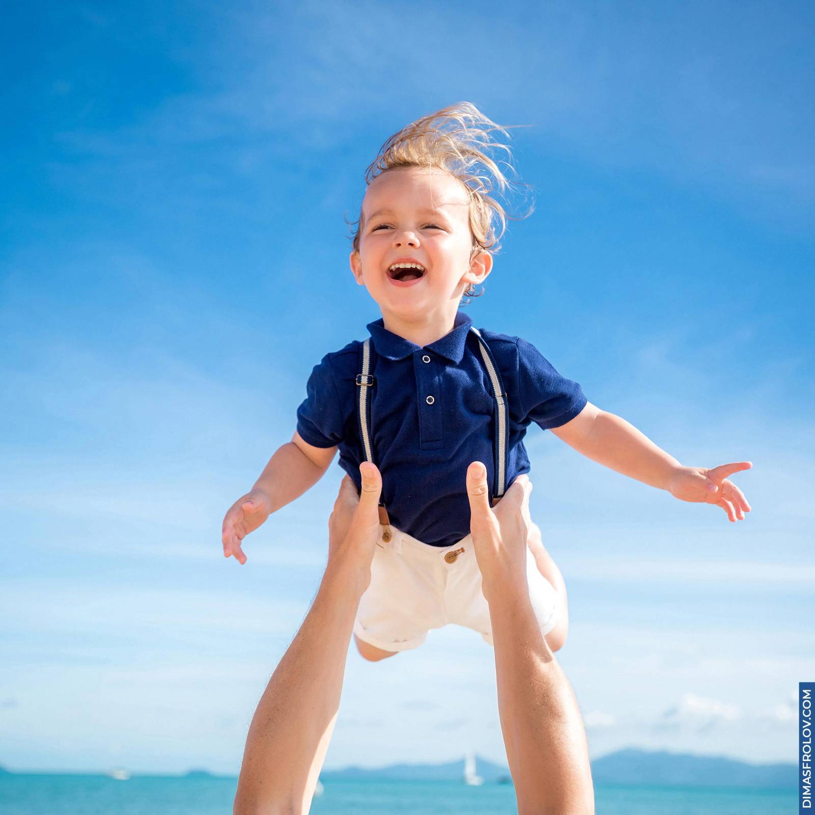 Happy toddler in suspenders being thrown into the air on the beach