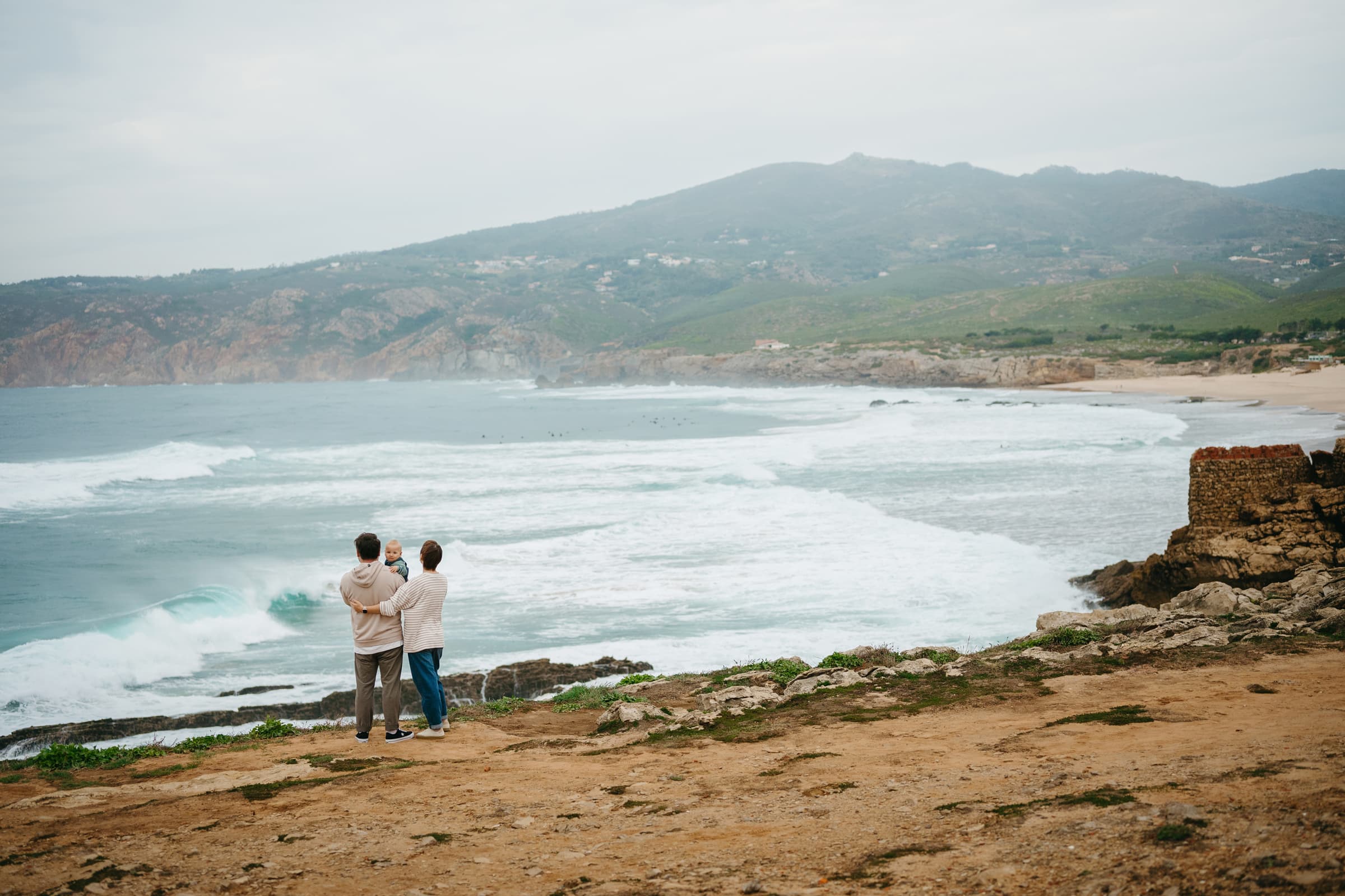 Parents holding a&nbsp;baby on&nbsp;the&nbsp;headland above Guincho beach with&nbsp;the&nbsp;Sintra hills behind, family photoshoot on&nbsp;the&nbsp;Atlantic coast