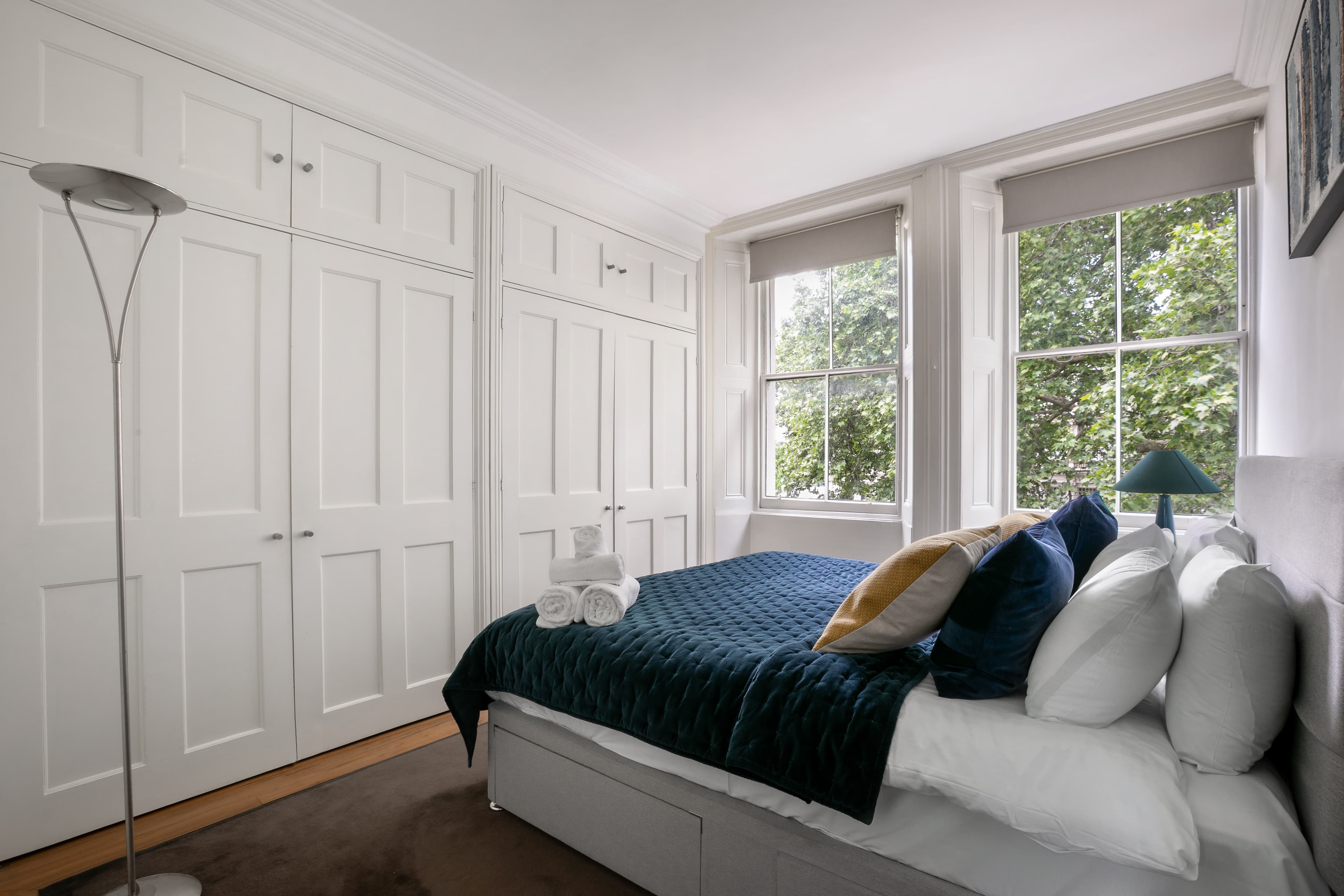 A bright bedroom with two windows showing greenery outside and a blue quilt on the bed